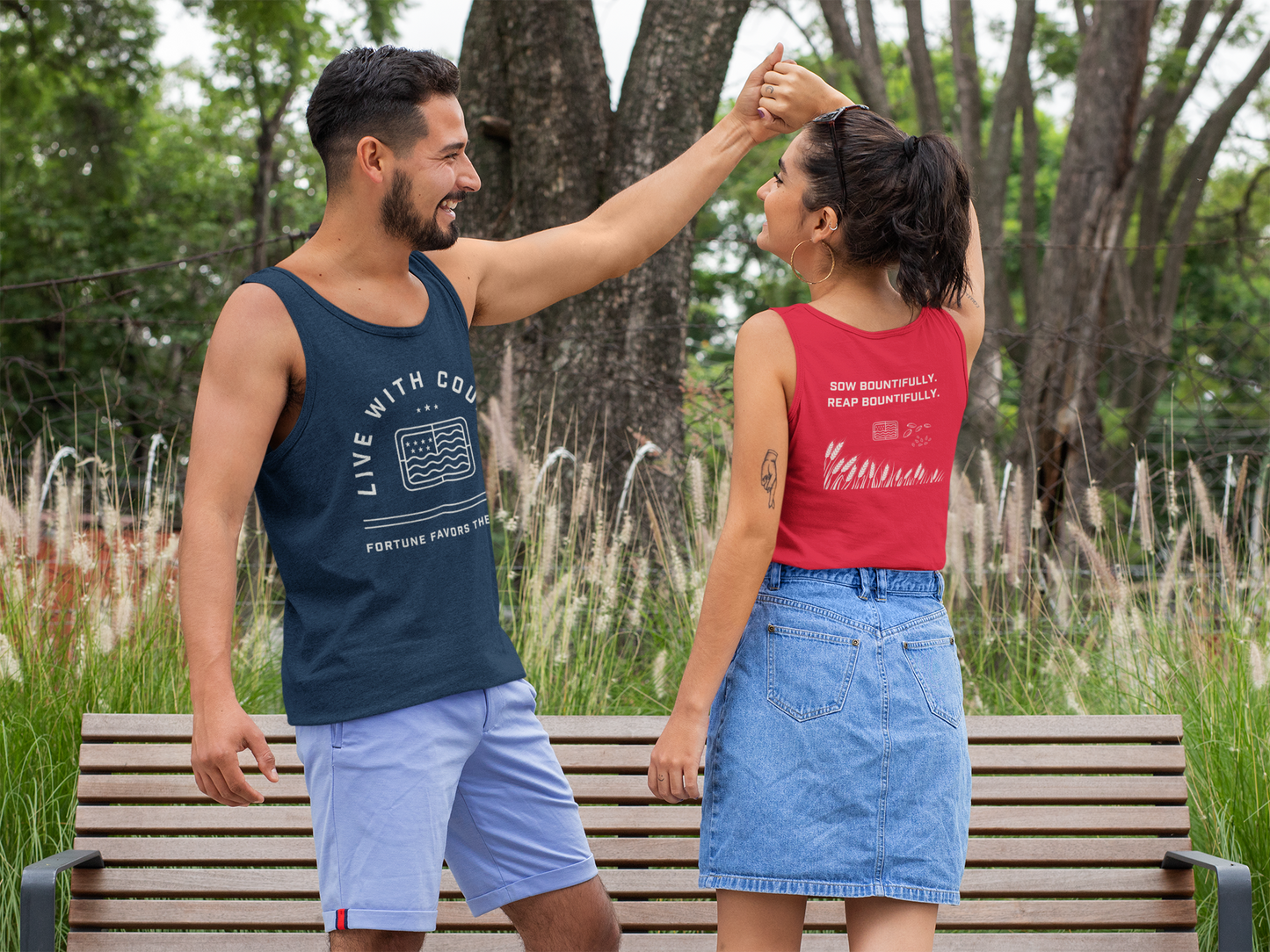 Man and woman dancing on a bench in a park with trees and grass in the background with Live With Courage tank tops.