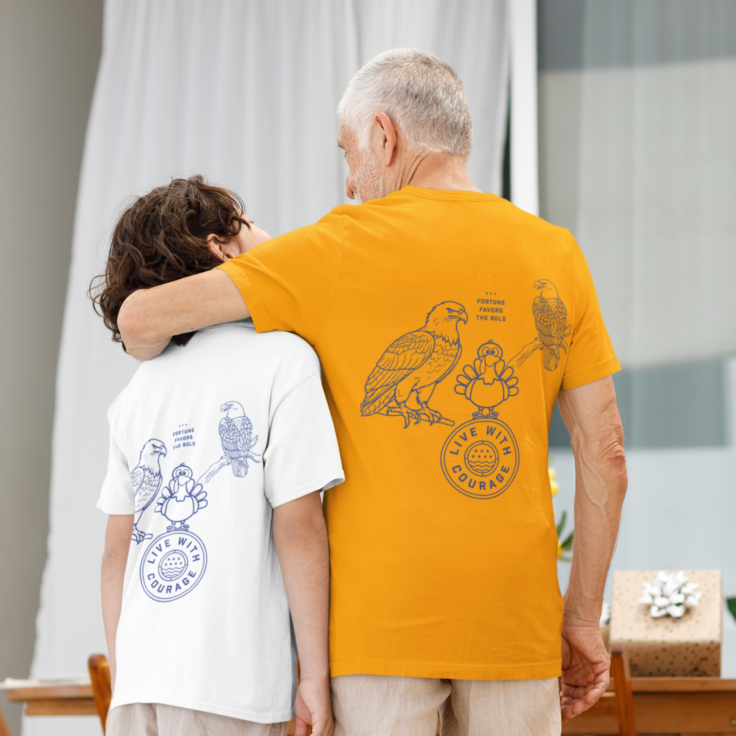 Grandfather and grandson stand arm in arm wearing matching Live With Courage® Thanksgiving t-shirts featuring a humorous turkey among eagles and the phrase 'Fortune Favors the Bold' on the back